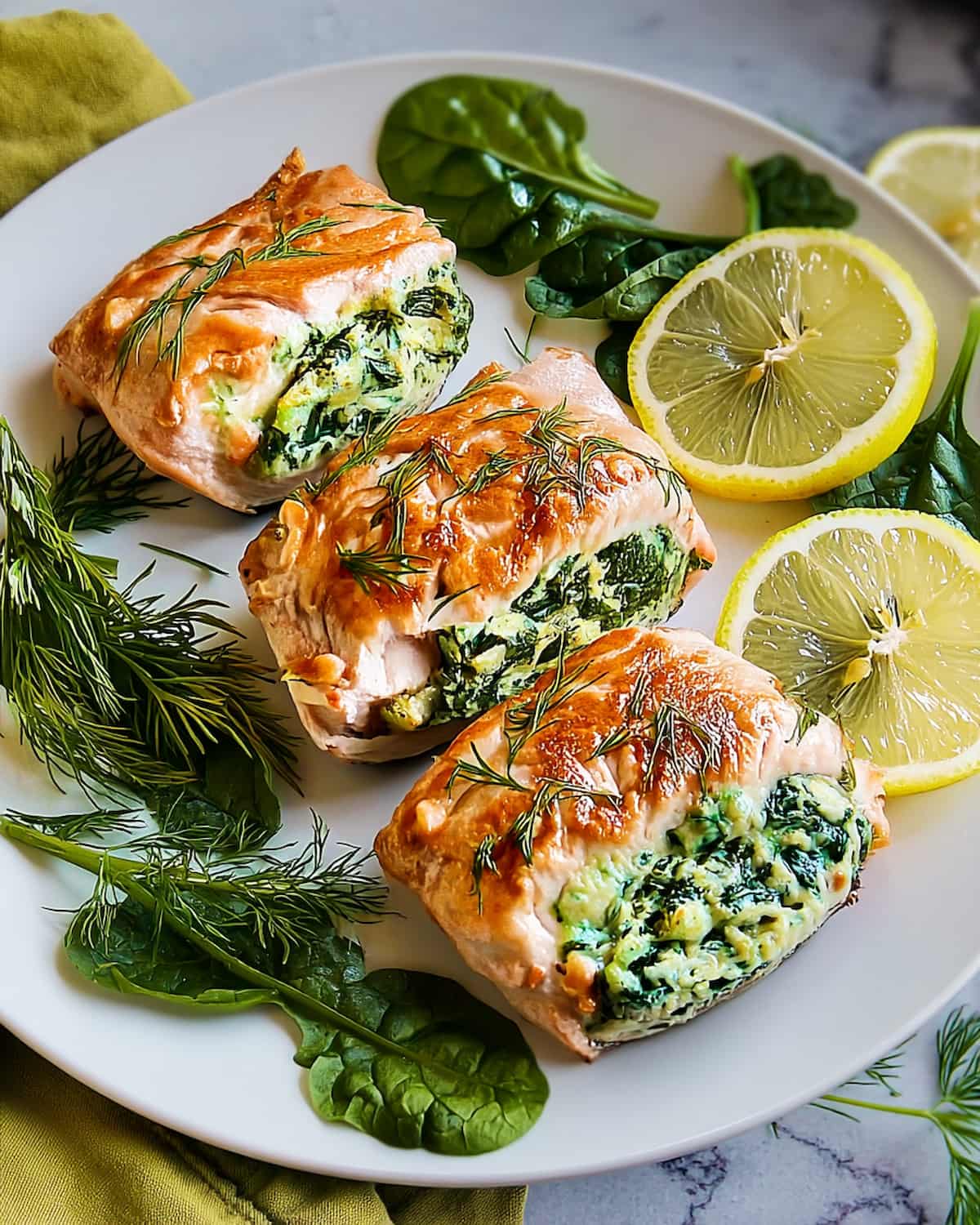 Spinach and cream cheese stuffed salmon fillets on white plate with lemon slices, fresh dill, and spinach leaves as garnish.
